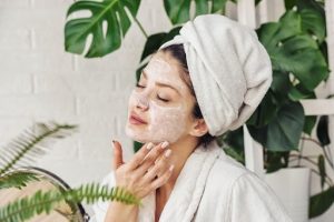 Woman with a towel wrap on her head exfoliating her face in front of plants