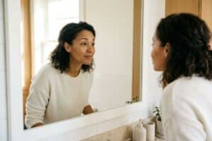 Adult in a cream sweater leans toward a bathroom mirror, studying their reflection beside a soap dispenser and small plant.